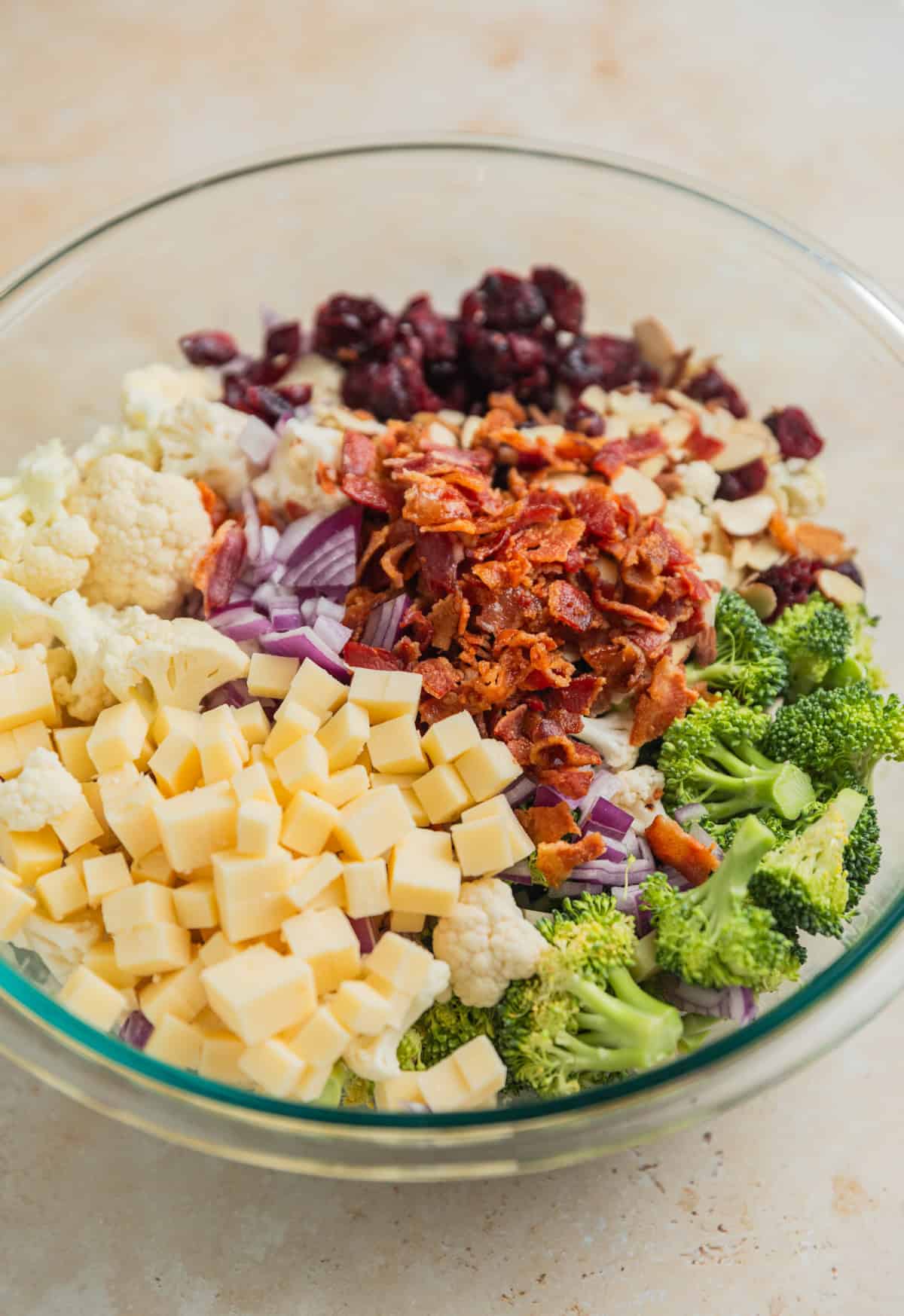 Bite sized broccoli, cauliflower, bacon, cubed cheese, and other ingredients arranged in glass bowl.