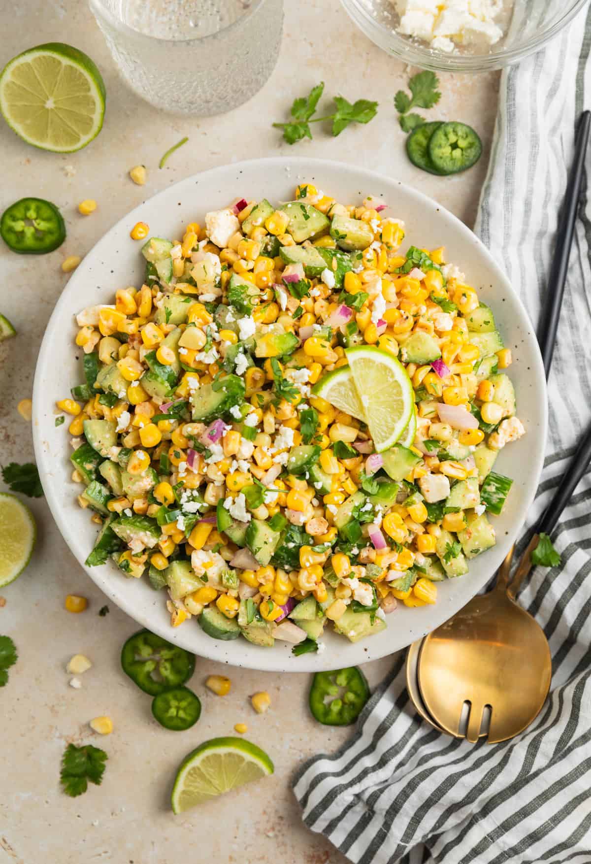 Overhead view of corn and cucumber salad in serving dish with lime wedges on top.