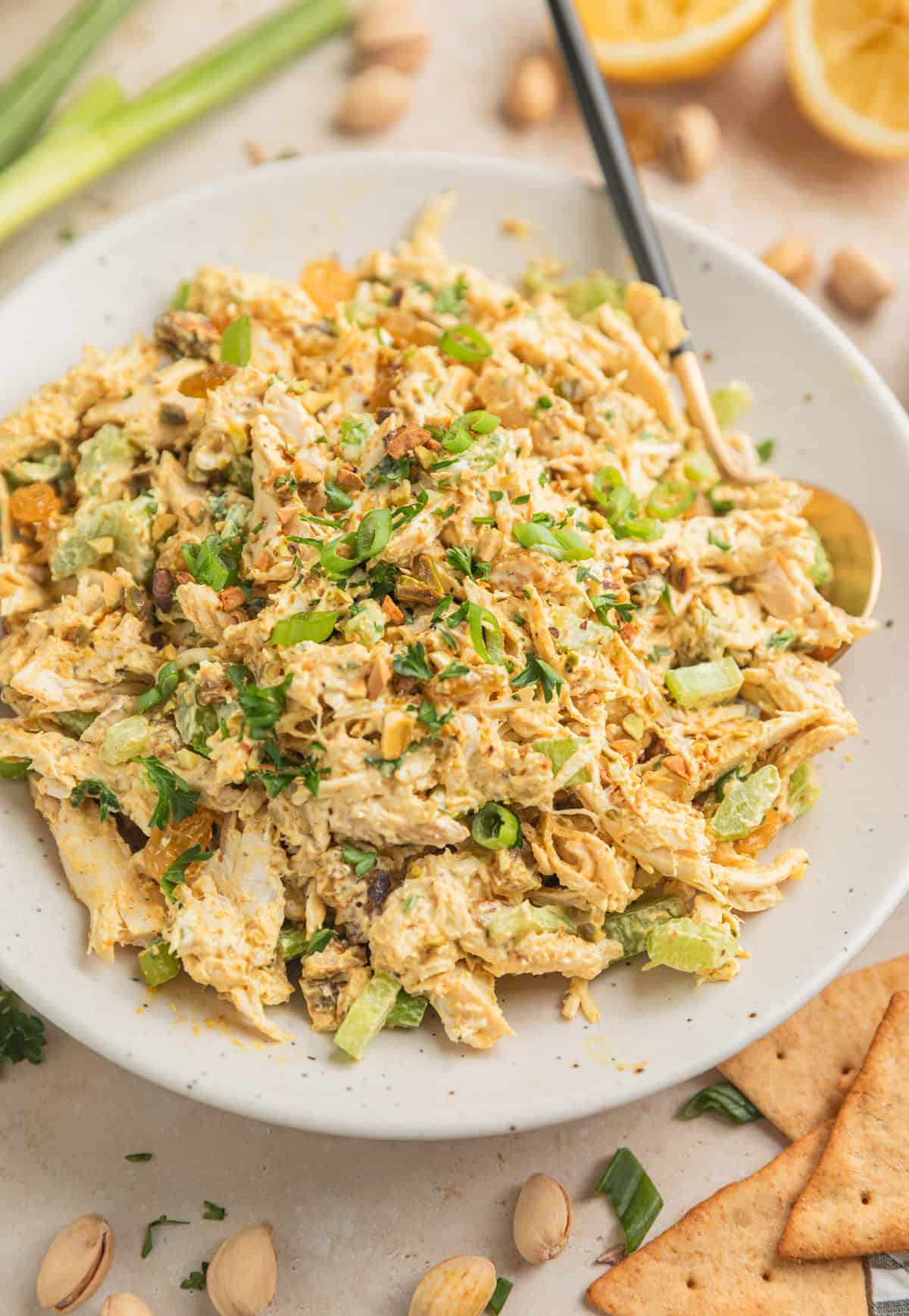 Curry chicken salad in dish with spoon topped with chopped green onion, chopped pistachios and parsley.