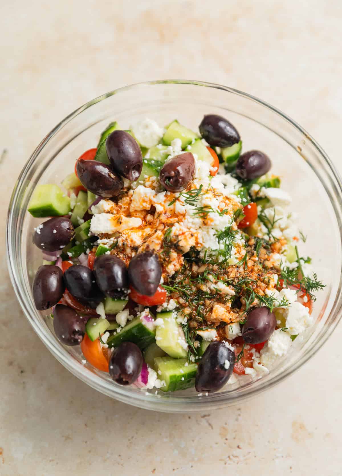Cucumbers, olives, tomatoes, feta and dressing in glass bowl before tossing.