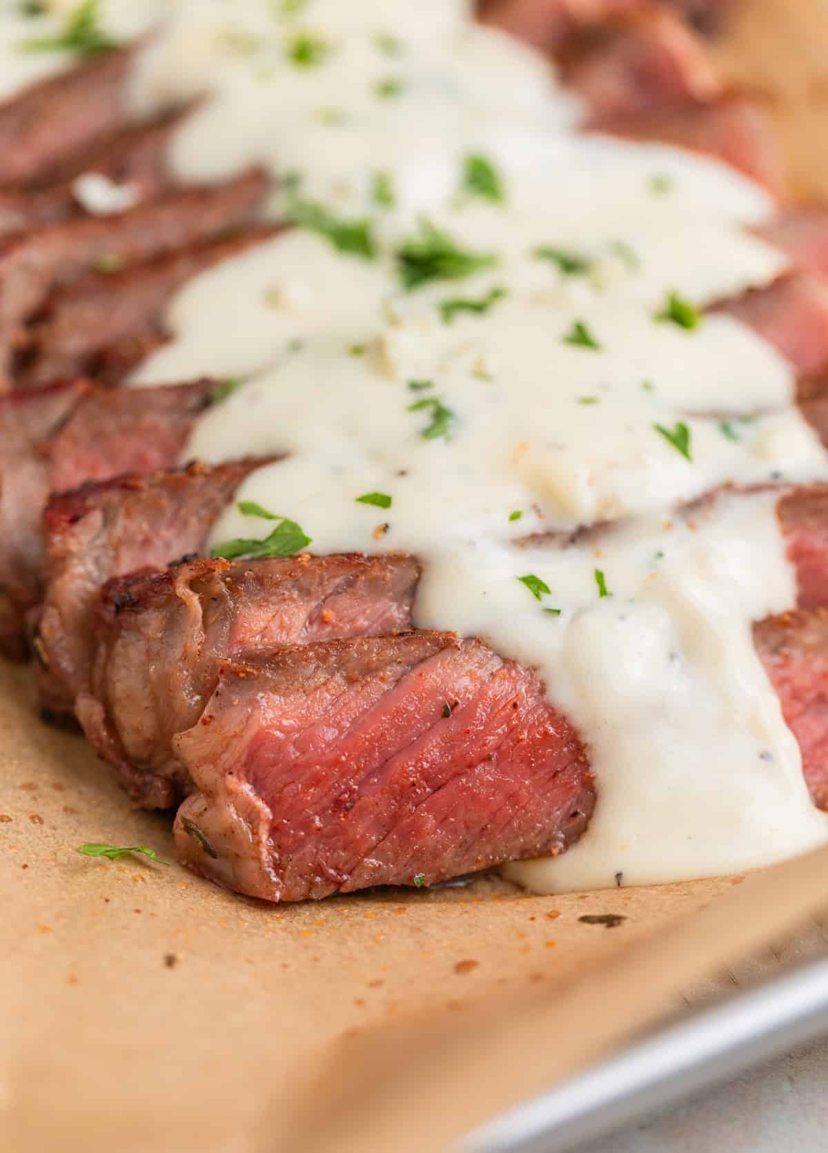 Sliced ribeye steak on parchment paper with cream sauce over top.