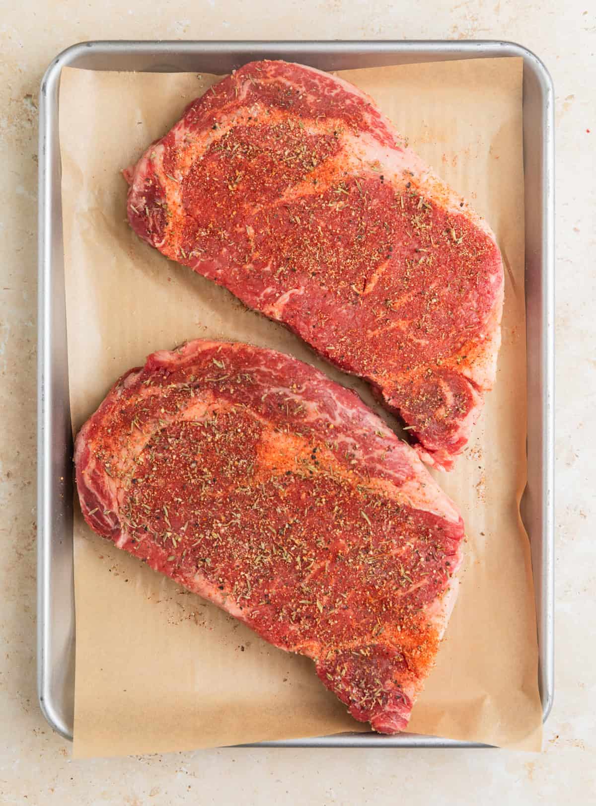 Seasoned ribeye steaks on parchment lined pan.
