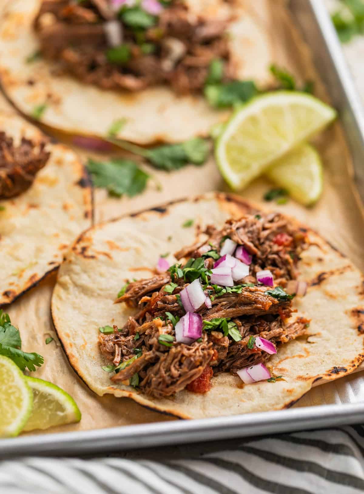 Shredded beef tacos on parchment lined pan topped with onion and cilantro.