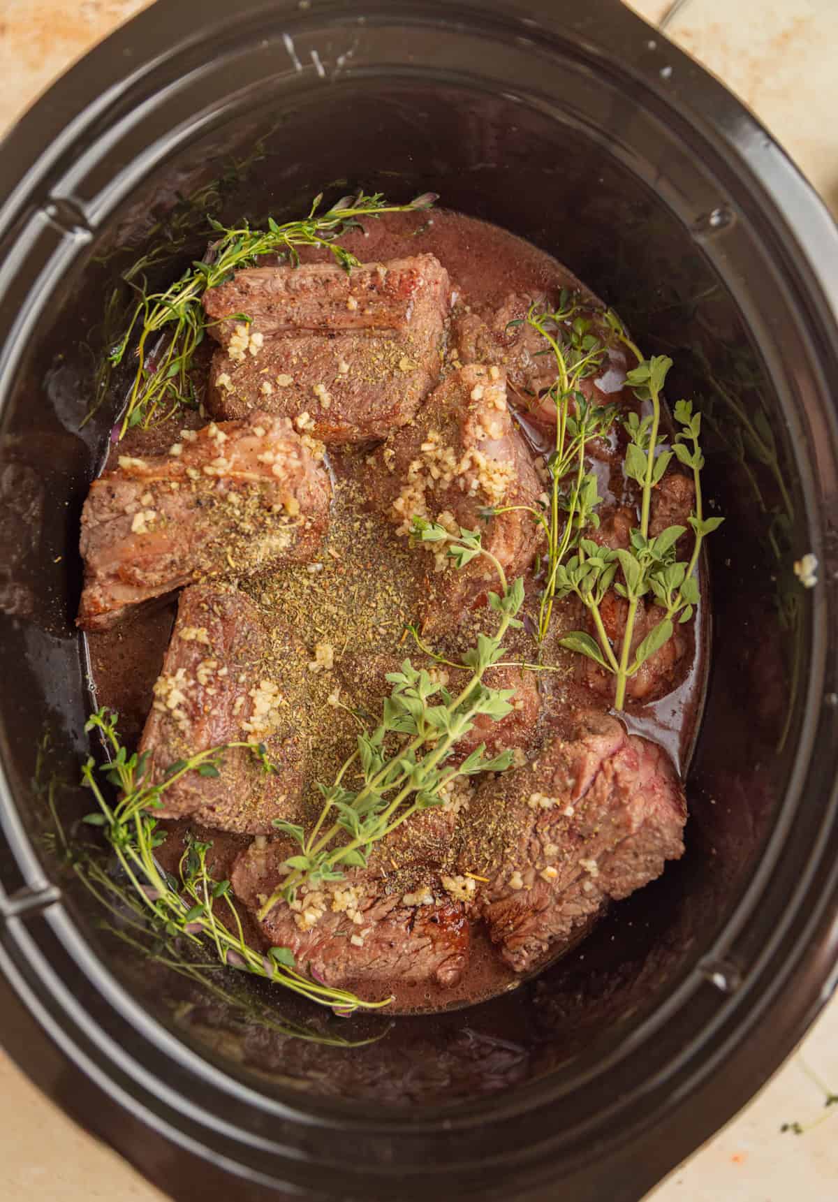 Slow cooker with short ribs, broth and herbs before slow cooking.