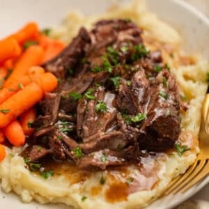 Short ribs with gravy over top on plate with potatoes and carrots.
