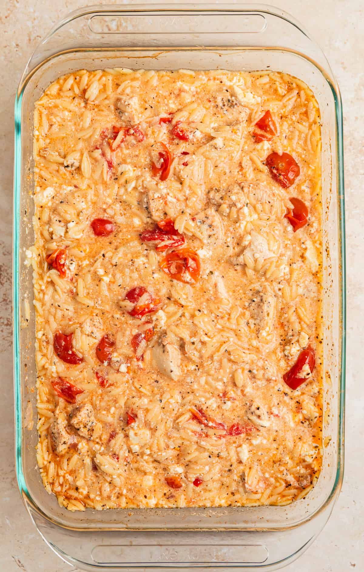 Feta mixed into Greek chicken and orzo bake in glass dish.