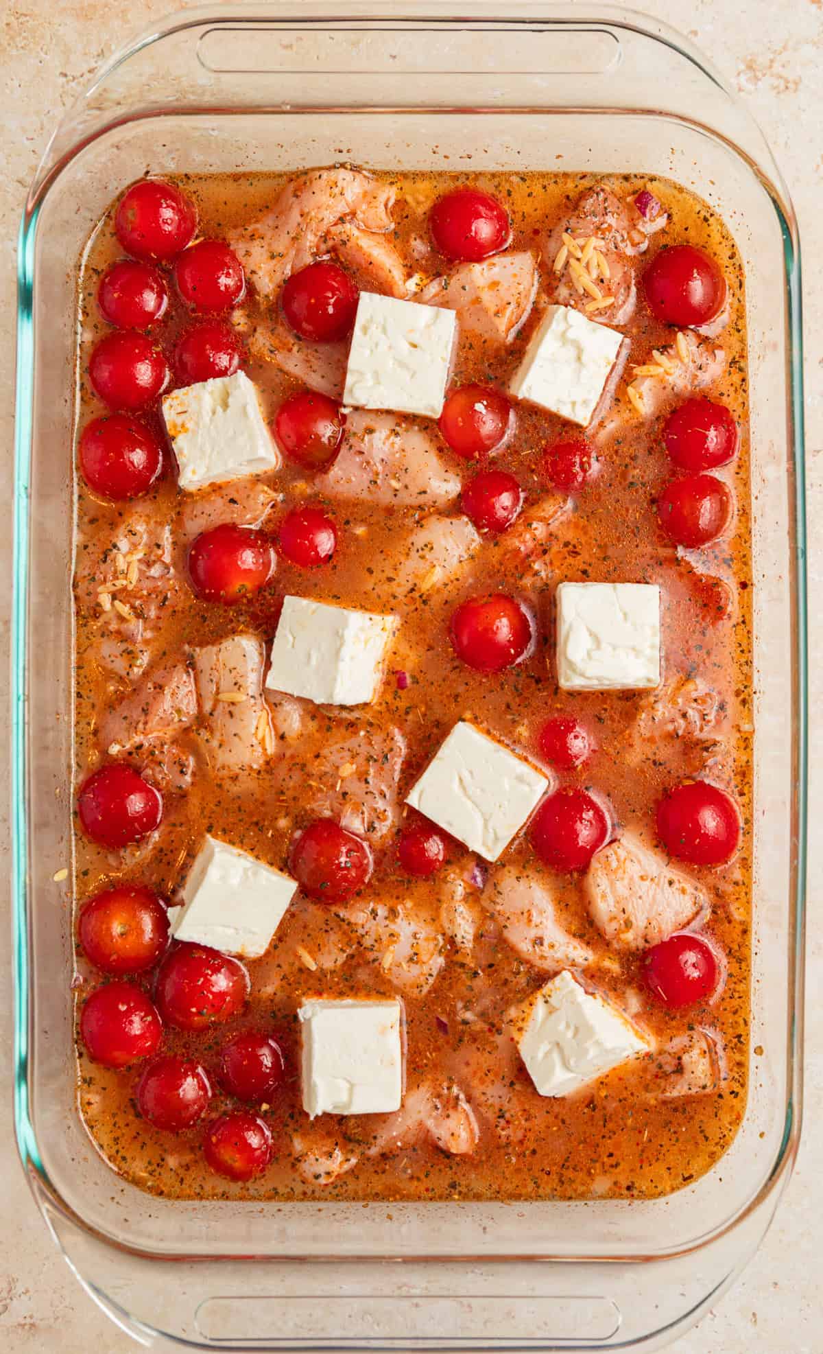 Chicken, orzo and tomatoes in baking pan with broth and feta cubes.