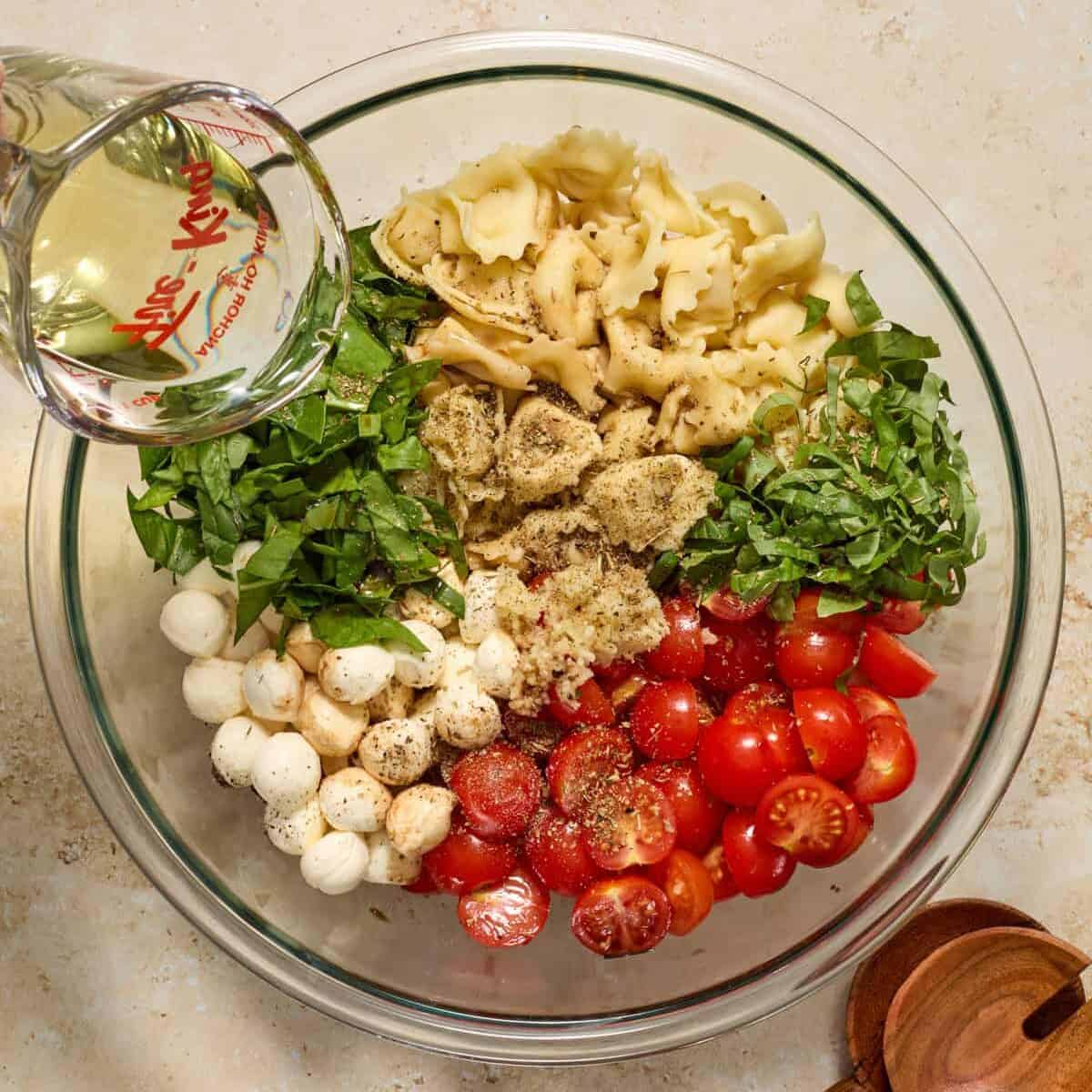 Pasta salad ingredients in bowl with oil being poured over.