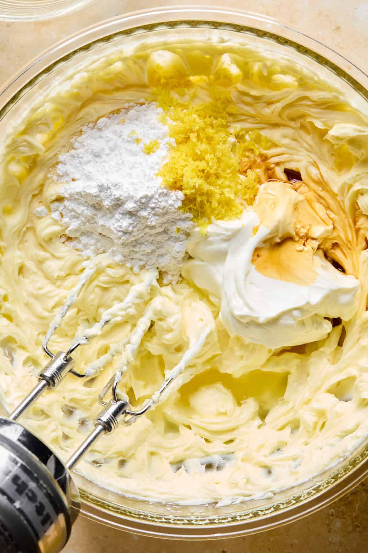 Sour cream, lemon zest, powdered sugar and other ingredients in glass mixing bowl with cream cheese mixture.
