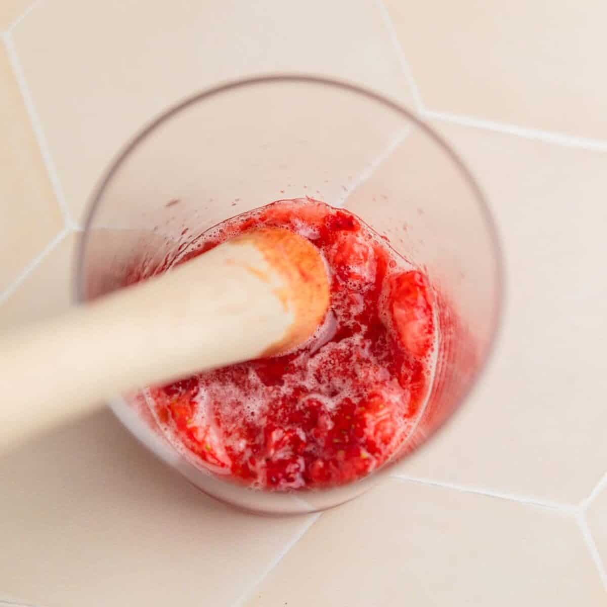 Muddles strawberries with maple syrup in glass cup.