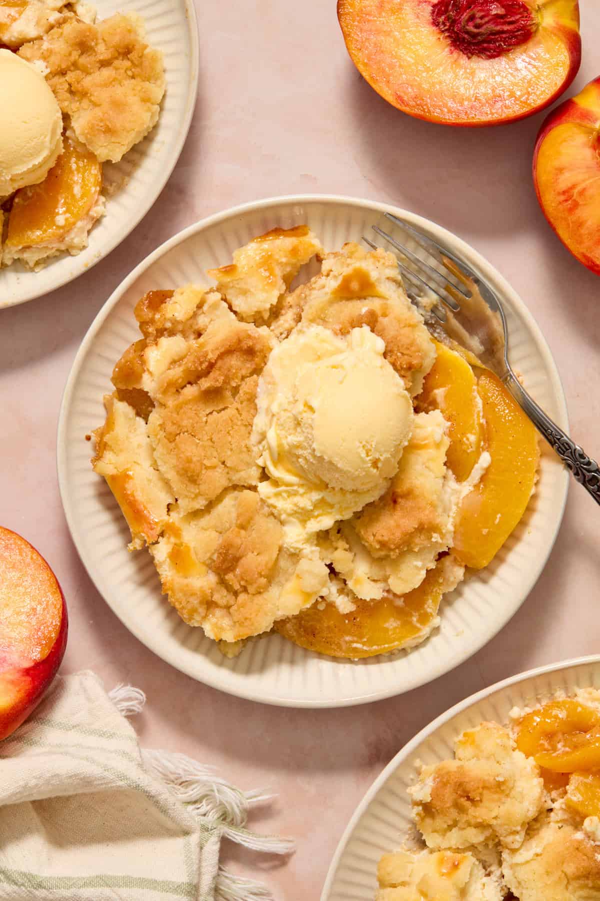 Cream cheese peach cobbler with scoop of vanilla ice cream on plate with fork and peaches surrounding.