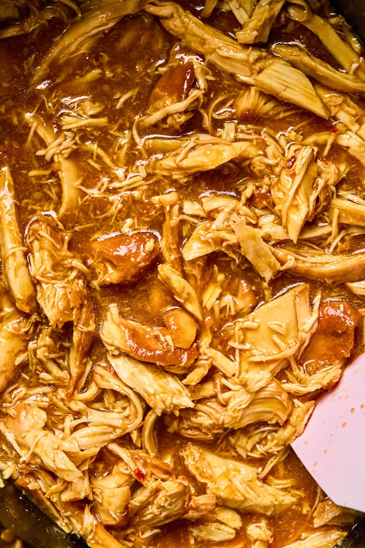 Close up view of slow cooker with shredded honey garlic and sriracha chicken with spoon.