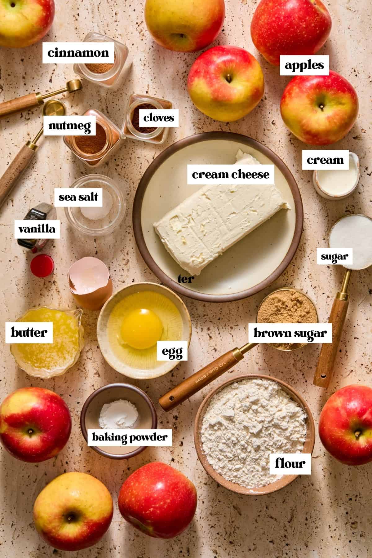 Cream cheese, apples, warm cpices, egg, flour and other labeled ingredients on surface.