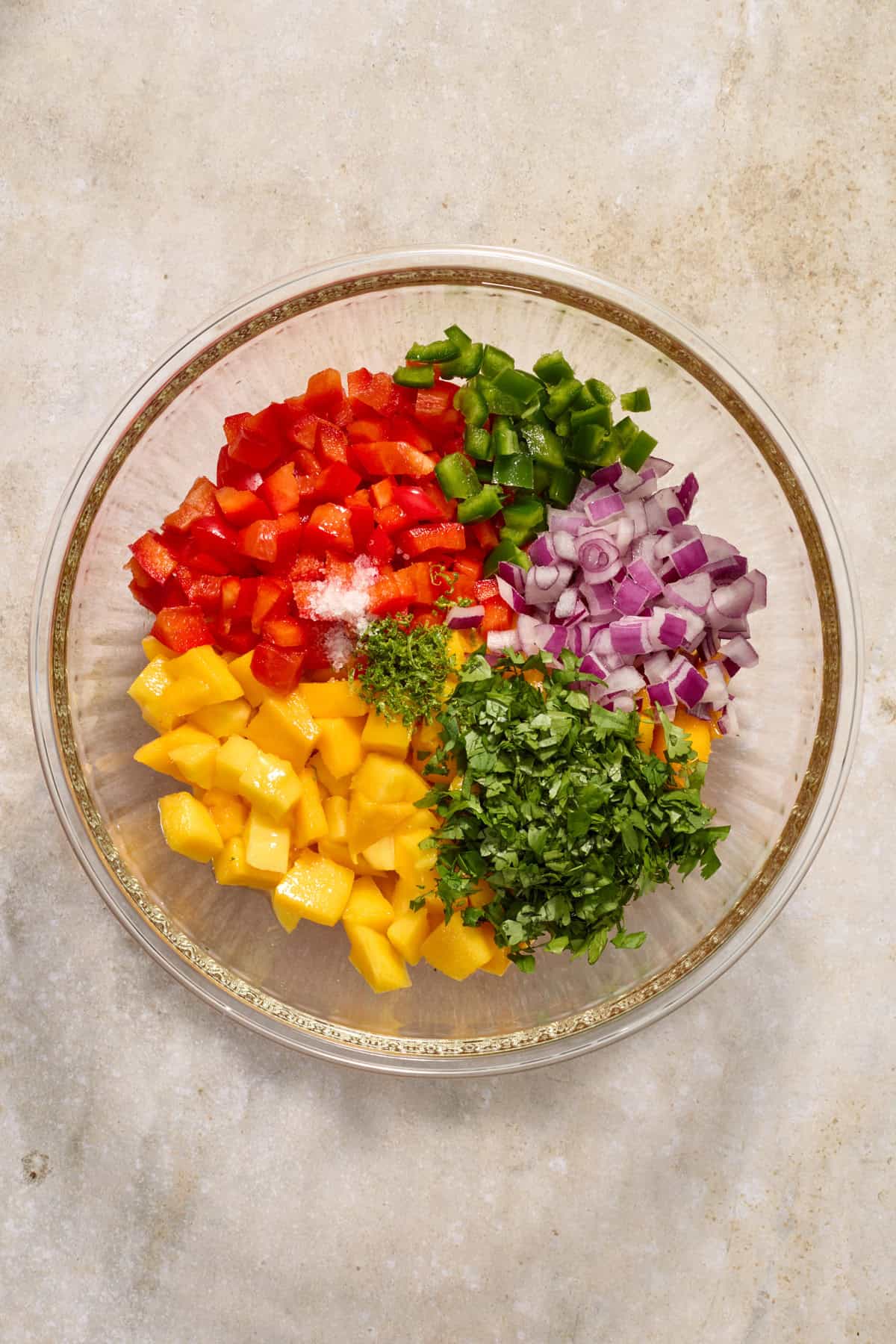Diced mango, onion, red bell pepper, cilantro and other ingredients in glass mixing bowl.