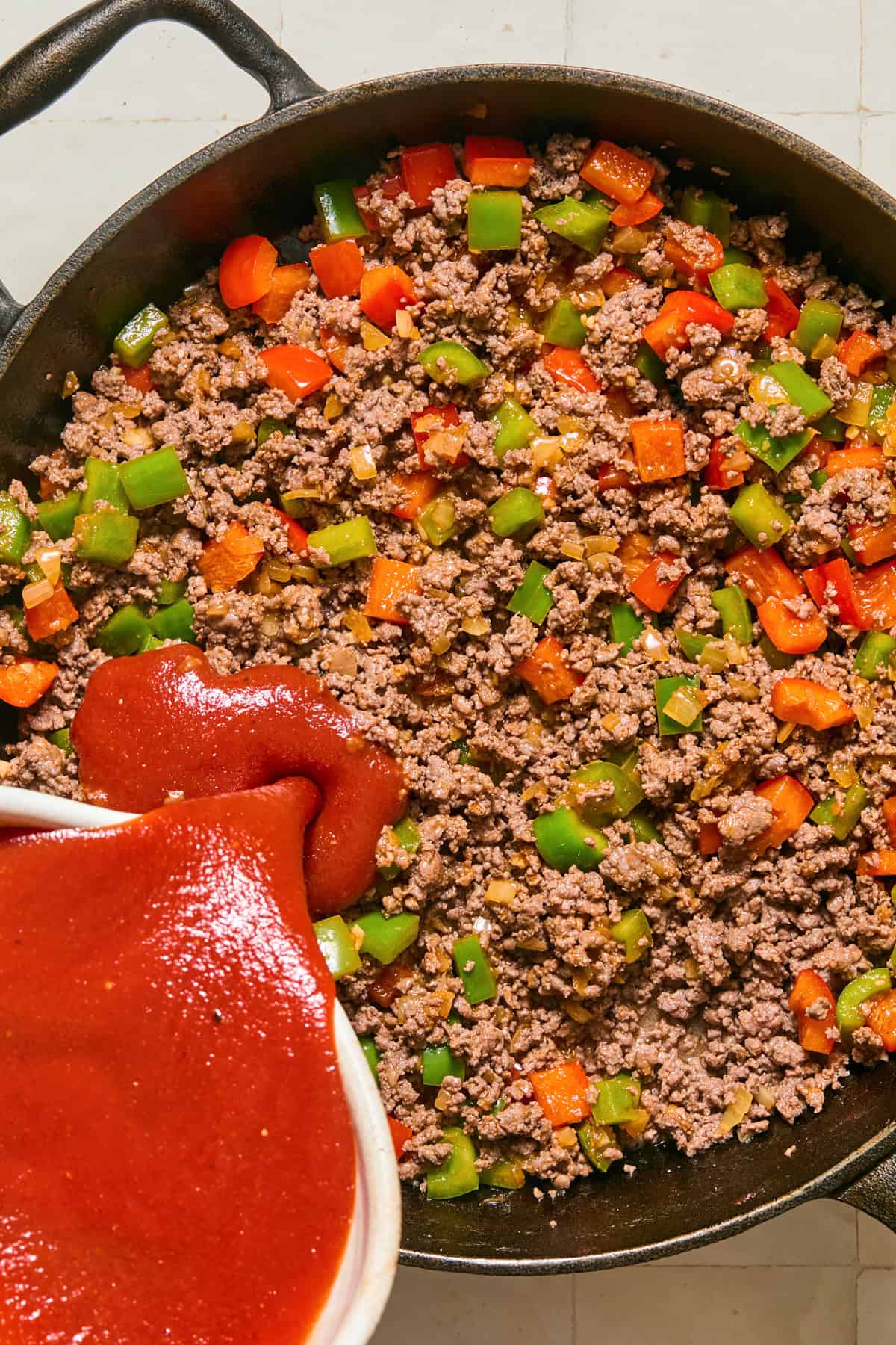 Browned ground beef and peppers with tomato mixture being poured in.