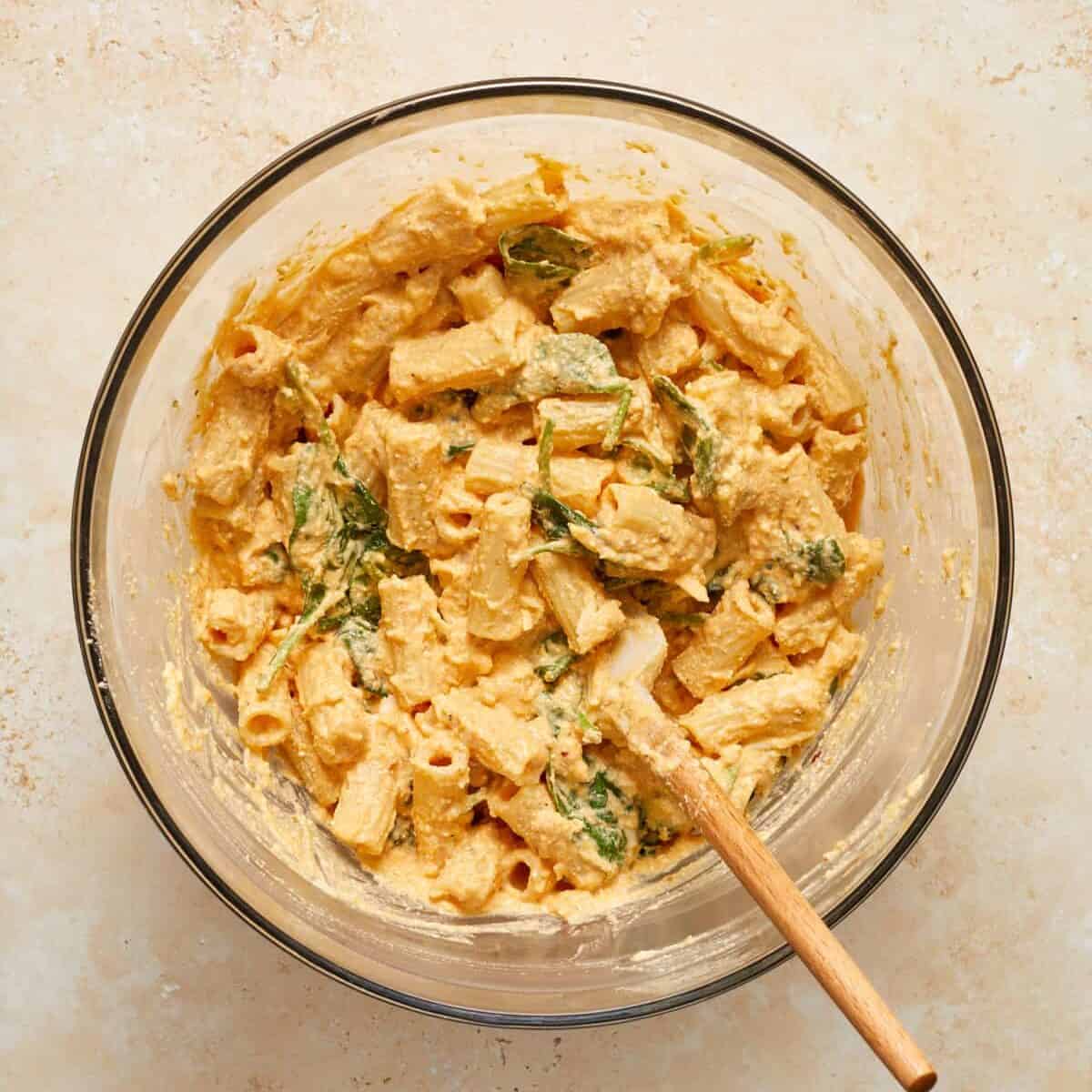 Pumpkin mixture tossed with pasta and spinach in glass mixing bowl with spatula.