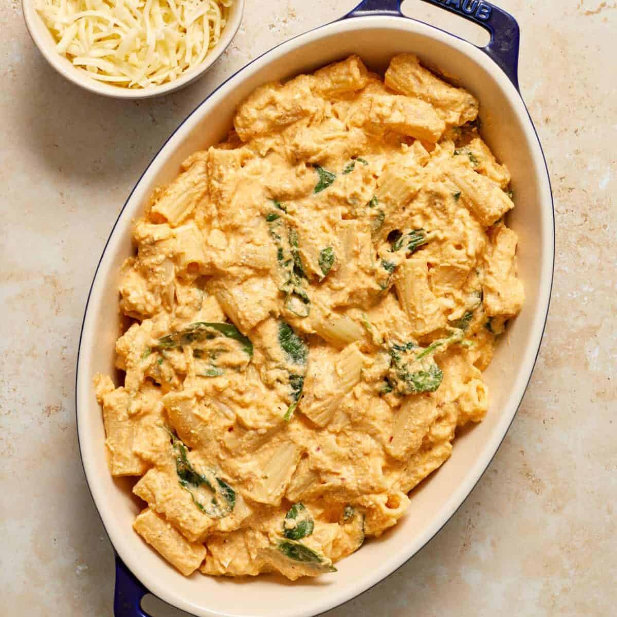 Pumpkin pasta bake in casserole dish with bowl of cheese beside it.